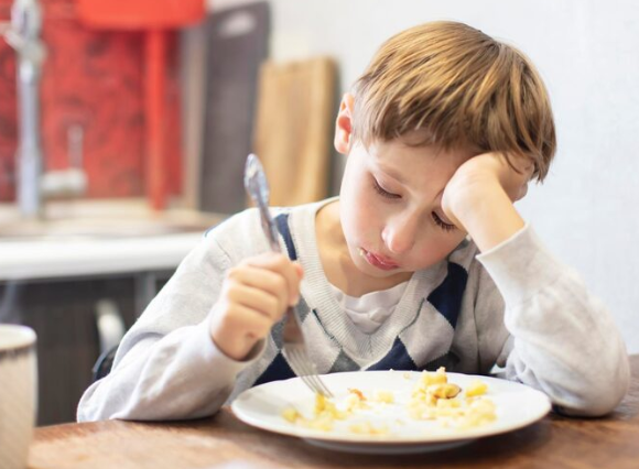 Child sitting at a kitchen table with a plate of food, holding a fork and resting head on one hand, appearing reluctant to eat.
